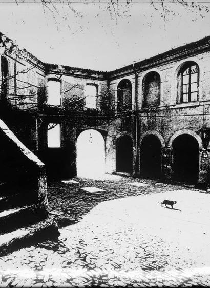 The image is a black and white photograph showing an old, dilapidated courtyard with crumbling walls and arched doorways. There are three large arches on the right side of the photo leading to darkened interiors, while two smaller windows can be seen above them. The ground appears uneven and cracked, covered in what looks like small stones or debris. A lone dog is walking down the center path towards a darker section at one end.
The sky is overcast with bare branches visible against it on the left side of the photo. There's an overall atmosphere of abandonment and decay. This particular image seems to capture a moment frozen in time, showcasing historical architecture that has seen better days.