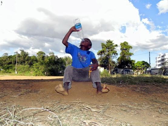 A young Malagasy boy, around 8 years old, sitting on a dirt mound, drinking water from a plastic bottle