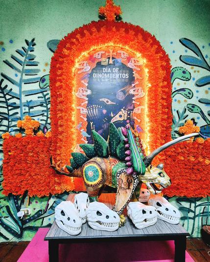 This photo shows a vibrant and colorful ofrenda, a traditional Mexican altar for Day of the Dead (Día de Muertos), honoring dinosaurs! There's a big, beautiful dinosaur with pointy plates on its back, surrounded by pretty orange flowers and smaller dinosaur heads.