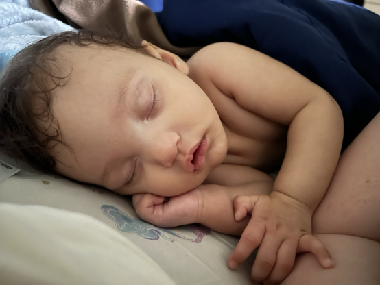 A light-skinned baby with dark curly hair sleeps with one hand curled under their face, and their other arm across their body resting on their elbow. Their mouth is slightly open in an expression of blissful innocence.