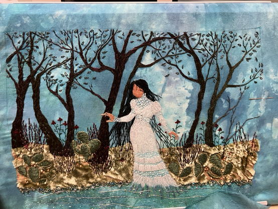 A mottle dyed blue muslin panel embroidered with La Llorona standing in the edge of a river, with cacti, wildflowers, and mesquite trees growing along the bank.