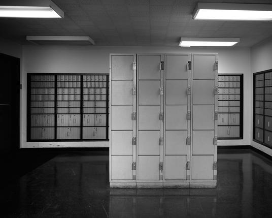 The image shows a room with rows of uniform mailboxes lining the walls. In the center, there is a large, rectangular structure also made of metal boxes with locks. The room is well-lit with fluorescent lights, and the floor is shiny, reflecting the light above.