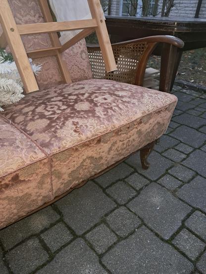 Sitzfläche eines Sofas. Die Armlehne ist aus massivem Holz, das mit einem Rattangeflecht an der Sitzfläche befestigt ist. Bezogen ist das Sofa mit einem lachsfarbenen Brokatbezug mit Blumenmotiven und OHNE Flecken oder Beschädigungen.