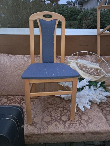 Auf dem Sofa steht ein Stuhl mit hoher Rückenlehne. Das Holz ist hell. Rücklehne und Sitzfläche sind mit einem blauen Stoff bezogen. Der Stuhl hat keine Beschädigung. Der Stoffbezug ist ohne Flecken oder Beschädigungen.
