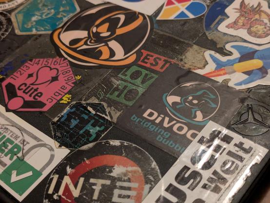sticker Bomb with some stickers looking really rough and others like new.
A Hexagonal cute seil, an alien in a bubble spacecraft, a bunny in a bubble spacecraft with divoc-logo, a dragon with a die, a polyamory relationship heart and a lot more sticker bomb.