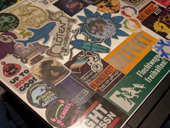 A close up of the second laptop. All the stickers are a lot newer, but a DEFECT sticker with silver printing on neon paper is already showing signs of age. A holographic Moon sots on top of a flower in the middle of the laptop. The moon was perfectly put on top of the flower.

Noticeable are also two stickers with similar theming for a fantasneak angel sticker with a hand forming a snake head and a B-Engel sticker that has a halo with horns. The stickers are matte and are printed on silver foil, making them reflect light with kind of an underglow.