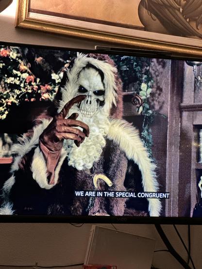 A television showing the Sky TV version of The Hogfather, based on the book by Sir Terry Pratchett.  On screen is Death, an anthropomorphic personification, a walking skeleton usually wearing a hooded black robe but instead wearing a red velvet robe trimmed in white fur. His hand is clothed in a leather glove, and he is gesturing with the index finger out.  He is in a wood paneled room with a decorated holiday tree in the background.  The closed captioning on the screen says “WE ARE IN A SPECIAL CONGRUENT”