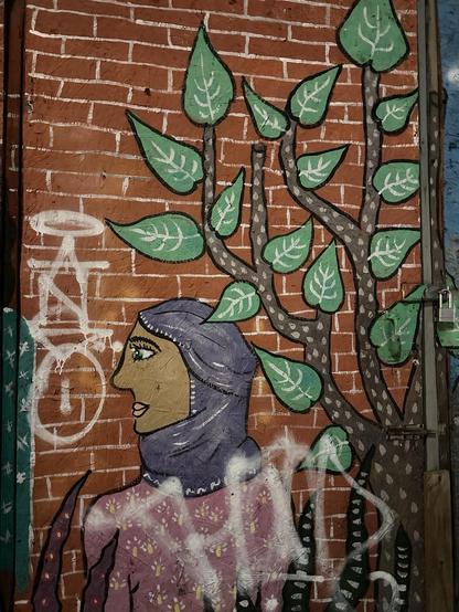 a mural painted on a brick wall depicting a woman wearing a purple hijab next to a leafy plant. she seems to be gazing at ANSO in vertical white spraypainted letters with a halo over the A. there is another white tag over her shoulders