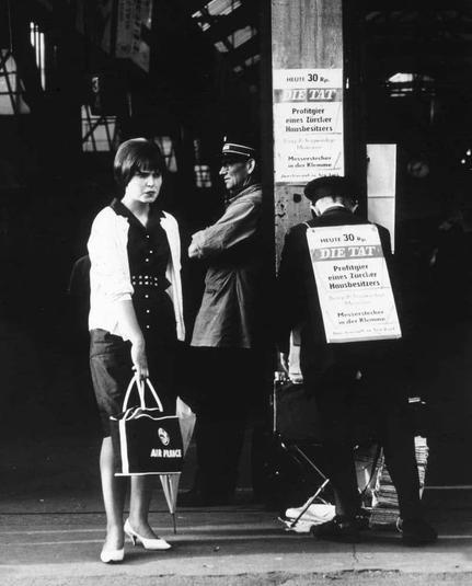 The image is a black and white photograph featuring three people on what appears to be an urban street scene, possibly from the mid-20th century. On the left stands a woman facing away from the camera; she has short hair, wearing a light-colored blouse with dark vertical stripes or accents at her waistline, paired with darker trousers. She is holding a purse and another bag that reads "AERO PLAZA." To the right of this figure are two men standing near a cart bearing several items including newspapers stacked on top; one has his back to us, wearing what seems like a uniform possibly related to railway or transit services. The other man stands beside him in profile view, looking toward something out of frame with an expression that could be interpreted as contemplative or attentive. In the background is signage for "DIE TAT," likely referring to newspapers from Germany's major tabloid publications during this era; it also includes text suggesting prices and services related to these papers. The setting has a gritty, atmospheric quality indicative of city life at the time depicted in the image.