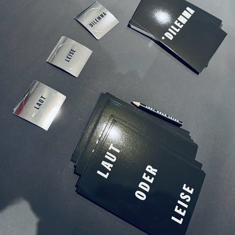 Blick auf einen dunklen Tisch mit zwei Stapeln schwarzen Postkarten, darauf in weiß zu lesen „LAUT ODER LEISE“ und „*DILEMMA“ Darüber drei kleine Stapel silberne spiegelnde Sticker mit den Worten „LAUT“, „LEISE“ und „DILEMMA“ in schwarzer Schrift.