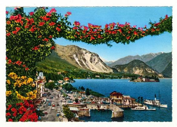 The image presents a vibrant panorama of Lago Maggiore, specifically focusing on the town of Stresa. It is framed by an overhead archway filled with lush greenery and blooming red flowers that add to its picturesque charm.

In the foreground, we see a bustling waterfront promenade lined with various buildings, cafes, restaurants, and shops catering to tourists. A large ferry docked at the pier indicates active transportation on the lake itself. The water is calm, reflecting the clear blue sky above.

The middle ground showcases Stresa's urban landscape featuring apartment buildings in pastel hues alongside a busy street filled with cars from an era suggesting mid-20th century. It adds a sense of historical depth and cultural richness to the scene.

In contrast, the background offers breathtaking natural beauty with majestic mountains that rise steeply on either side of Lago Maggiore. Their rugged terrain provides a striking backdrop against the serene lake's tranquil waters. Nestled within these mountains is Isola Bella, an island resort known for its well-preserved gardens and palaces.

The overall composition captures Stresa as both a vibrant tourist destination and a harmonious blend with nature’s splendor, encapsulating the charm of Lake Maggiore itself. The image provides a glimpse into what life might have been like in this beautiful town during that period.