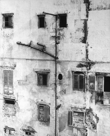 The image is a black and white photograph of the exterior wall of an aged, deteriorating building. The facade shows signs of wear with peeling paint and exposed brickwork in various states of decay. Multiple windows are visible; some have shutters closed while others appear boarded up or missing their glass panes entirely. A series of vertical pipes runs along one side of the structure, adding to its worn appearance.

The photograph is characterized by a stark contrast between light and shadow, emphasizing the textures and imperfections on the building's surface. The composition seems deliberate, capturing both the architectural details and the overall sense of neglect or abandonment. This image evokes a feeling of history and decay, possibly representing an urban environment in need of restoration.

The photo appears to be from around 1960 as indicated by the reference information provided: Bevilacqua, Carlo - Inabitabile (Inhabitable), circa 1960. The source link suggests that this image may have been published or cataloged on a website called "Rivista Ferrania."