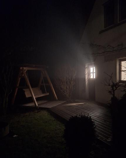 Eine Veranda mit einer Sitz Schaukel in der Nacht. Aus dem Gebäude strahlt Licht, dessen Strahlen in der feuchten Luft sichtbar werden.