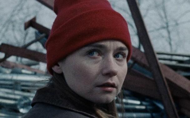 Jessica Barden wears a red stocking cap. She looks over her shoulder. Behind her are metal beams stacked haphazardly