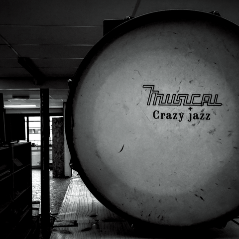 Black and white photo of a children's bass drum which is resting on a wooden shelf in a charity shop. Most of the drum is visible on the right of the frame. On the drumskin are the words: Musical & Crazy Jazz. Light is shining through the drum skin from the opposite side creating a circle.