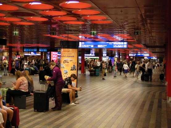 The inside of the Prague main train station entrance hall. The ceiling has some circular red light shades. The station is busy and full of passengers.