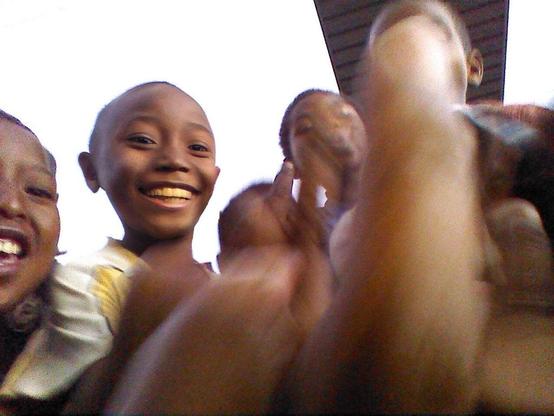 A playful and candid photo of five mischievous Malagasy boys, all approximately 10 years old or younger, crowded close to the camera with big smiles. Each boy is flipping the bird, and one hand with an extended middle finger is prominently in front of the camera, occupying nearly half the frame and out of focus.