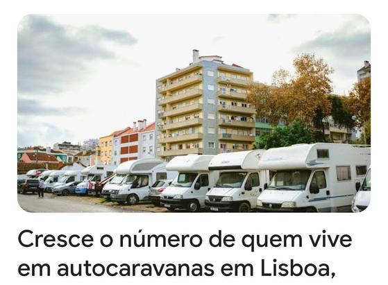 A foto mostra várias autocaravanas estacionadas em fila numa rua de Lisboa. Os veículos são brancos e estão estacionados defronte de prédios de apartamentos de vários andares. Há árvores com folhas amareladas ao fundo, sugerindo que a foto foi tirada no outono. A legenda da imagem afirma que o número de pessoas que vivem em autocaravanas em Lisboa está aumentando.