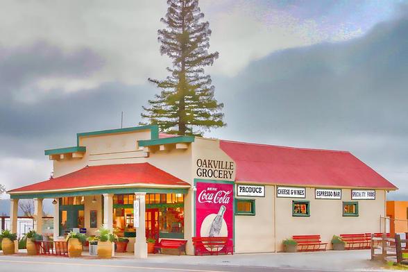 Oakville Grocery

On the side of Highway 29 in beautiful Napa Valley is the fabulous Oakville Grocery store. This landmark store dates back to 1874.
The front of the store has a porch supported by wood pillars.
In front of the porch are four wine barrels being used as planters have nice green plants growing in them.
The store is painted with a very light brown/tan with green trim. The roof is a red tile roof.
On the side of the store is a large Coca-Cola advertisement just below the Oakville Grocery sign.
Above the store you can see a tall redwood tree that is on the other side of the store.