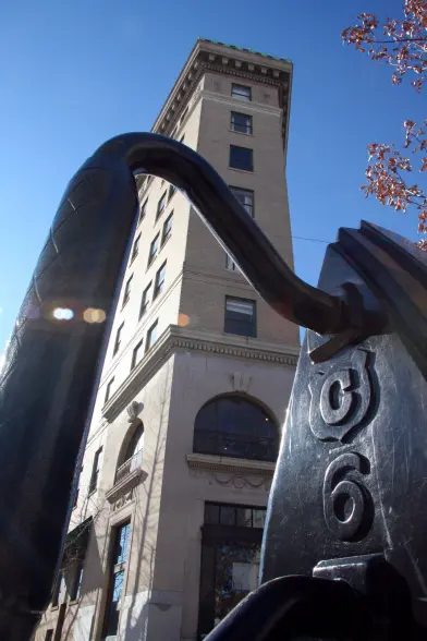 The image shows a tall, flatiron-shaped building with a distinctive tower. The building appears to be constructed of brick or stone, with detailed architectural features like arched windows and intricate decorative elements. In the foreground, there is a large, antique-style flat iron sculpture that complements the shape of the building. The flat iron sculpture has a prominent, sweeping curved form that extends out from the base, creating an interesting visual interplay with the angular, tapered facade of the flatiron building. The overall scene suggests an urban setting, with a clear blue sky visible in the background. The lighting and angle of the shot create an engaging interplay of shadows and highlights on both the building and the sculptural element.