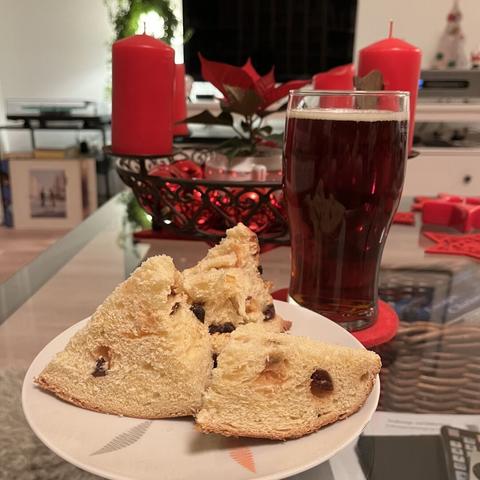 Ein Glastisch, darauf ein Teller mit großen Panetone-Stückchen. Daneben ein Glas mit dunklem Bier. Dahinter ein Adventskranz mit vier roten Kerzen