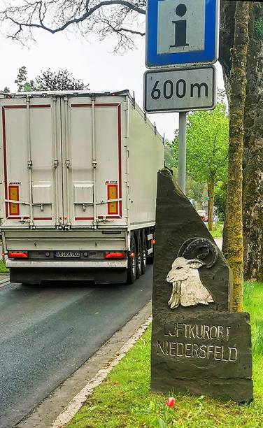 Links: Ein LKW fährt in den Ort Niedersfeld ein. Rechts oben ein blaues Schild mit dem Symbol "I" für Information, darunter 600 m. Rechts unten ein Stein mit dem Ortswappen: ein Ziegenbock - Brustbild, Profil.