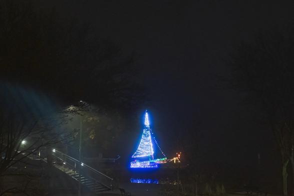 A sailboat brightly lit with Christmas lights and a reindeer made with Christmas lights, under the fog along the Charles River, with faint reflections visible. Surrounding trees are silhouetted against the night sky, and the scene is enhanced by soft illumination from nearby lamps.