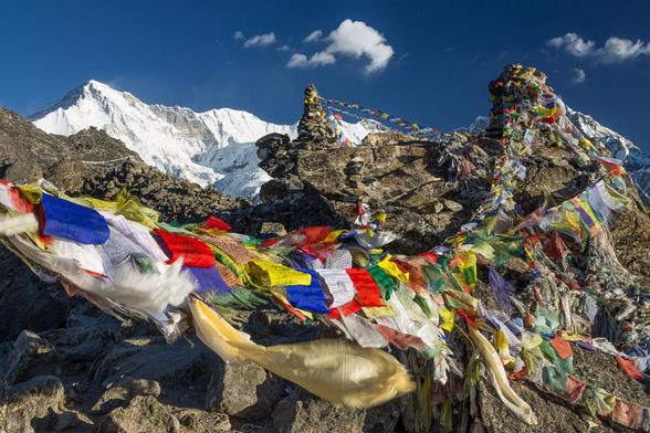 A colour photograph of a mountain summit decorated with Buddhist prayer flags seen on a clear, sunny day under the blue sky with a snow capped mountain in the distance.