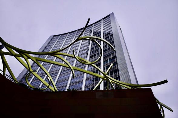Photograph looking up at the old AMP building in Sydney.  The building is a tower of steel, glass and white/grey tiles against a monotone grey sky.  In the foreground is a green spiralling metal sculpture.