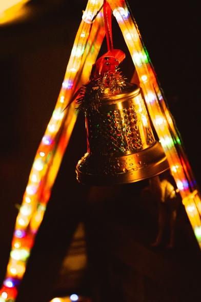 A glowing golden bell ornament hangs elegantly, adorned with red ribbon and tinsel, illuminated by vibrant, multicolored string lights in the background, creating a warm and festive ambiance.