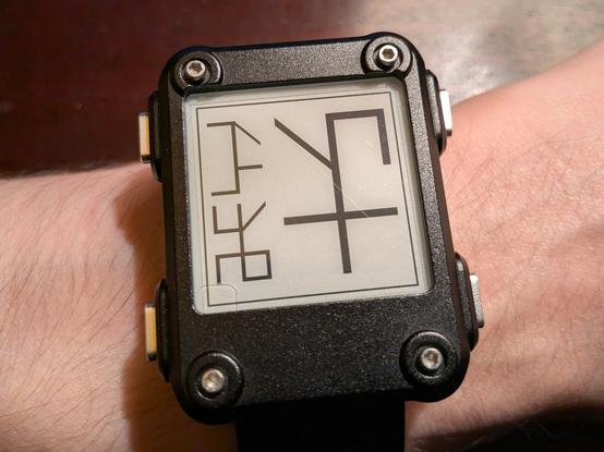 A photograph of a Watchy - an e-paper smartwatch - with three Cistercian numerals on screen representing time, date, and step count. A thin border runs around the edge of the screen.