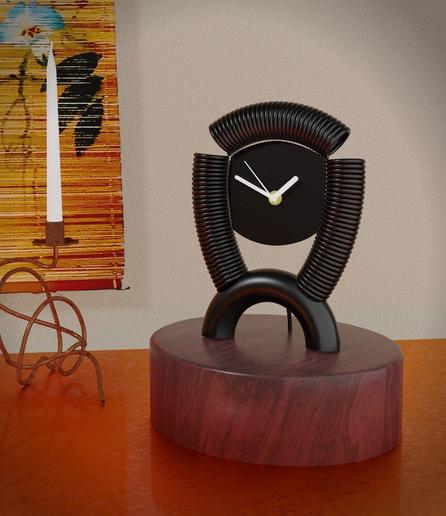 Auf einem dunklem Holzblock steht eine schwarze Uhr mit weißen Zeigern, um das Zifferblatt ist eine Spiralfeder gebogen und auf einen 180°-Rohrbogen geschweißt. Daneben ist noch ein Kerzenleuchter mit einer weißen Kerze aus verknotetem Kupferrohr und an der Wand dahinter noch ein Teil eines kakejiku (japanisches Rollbild) mit blauweißen Blütenmotiven zu sehen.

A black clock with white hands stands on a dark wooden block; a spiral spring is bent around the clock face and welded onto a 180° pipe bend. Next to it is a candlestick with a white candle made of knotted copper tubing and, on the wall behind it, part of a kakejiku (Japanese scroll painting) with blue and white flower motifs.