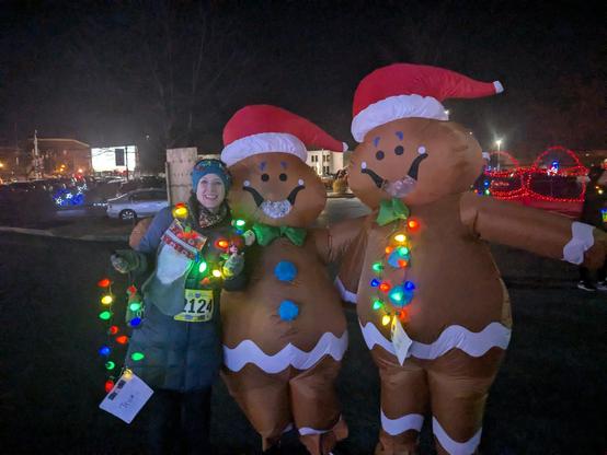 1 woman in a blue coat and 2 people in inflatable gingerbread costumes, all with Christmas light necklaces. Night time.