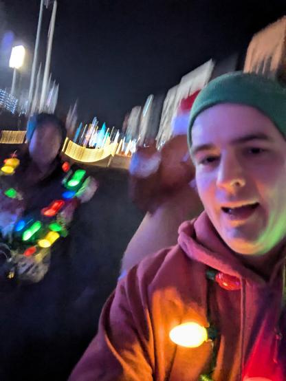 A blurry photo taken while jogging. A man takes the selfie on the right in a red sweatshirt and green hat. A gingerbread person and a woman are in the background. All have Christmas light necklaces. Stadium lights in the background