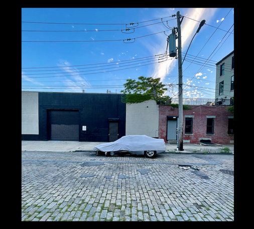 Une rue vide. Dans le ciel des câbles électriques reliés à un poteau. Vne voiture en stationnement est couverte d’une bâche de protection.