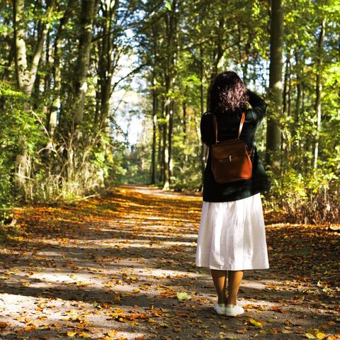 A lady in a green coat, long white skirt and with a brown leather backpack, taking a photo of a sunny path in the woods. The colors are a clear give-away of the season (autumn colors).