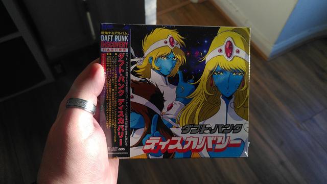 A photo of the Interstella 5555 limited edition of Daft Punk's Discovery album, in its protective film. The front cover is the album's original Japanese cover featuring the 4 blue alien characters from the animated movie Interstella 5555 with Japanese writing and a small sleeve on the left hand side.