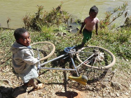 Two young Malagasy children, around 3 years old, are playing with an adult-sized bike lying on its side near a lake. The children appear slightly apprehensive about the photograph