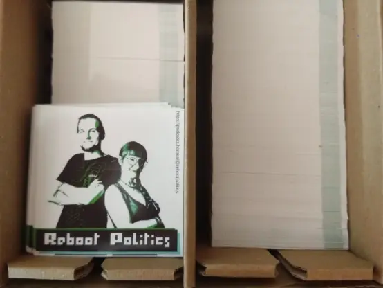 Zwei liegende Stickerstapel in einem Pappkarton. Auf dem linken Stapel sind einige Sticker nach oben gedreht, sodass die Vorderseite nach oben zeigt. Darauf 2 Personen, Flo und ich, Schulter an Schulter mit verschränkten Armen, darunter in Pixelschrift der Schriftzug "Reboit Politics". Der Sticker ist in weiß, schwarz, türkis und grün gehalten mit kräftigen hell-dunkel Kontrast.