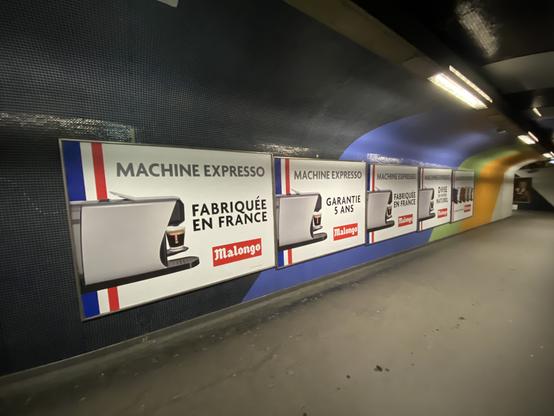 Couloir du métro de Paris, carrelage au mur, et une enfilade de cinq panneaux publicitaires pour la « machine expresso fabriquée en France Malongo ».