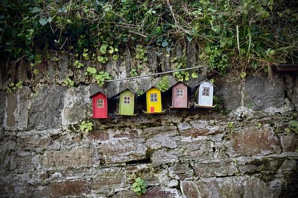 A row of small, colorful birdhouses—red, green, yellow, pink, and blue—is mounted against a stone wall, surrounded by greenery.