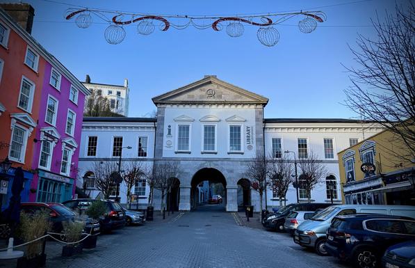 A view of a square in Cobh, Co Cork  featuring a building with an arched entrance, flanked by colorful buildings in shades of pink, orange, and yellow. The scene includes parked cars and decorative lights overhead, set against a clear blue sky.