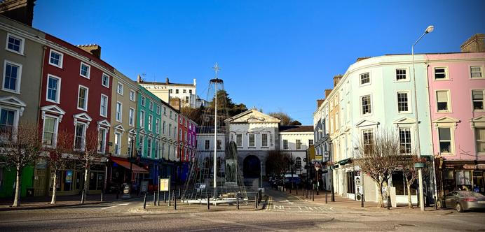A square featuring colorful buildings in Cobh Co Cork. The architecture includes a mix of pastel shades, and the sky is clear and blue. Trees with bare branches line the street.