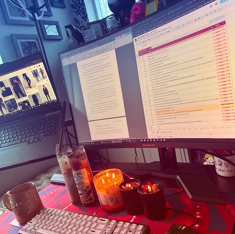 Rainy day moody picture of my desk. You can see 2 monitors, one with Pinterest open to some gothy clothing and one with a draft and a revision spreadsheet. The desk has 5 dramatic lit candles on it, a few for good sales and a few with lots of flowers and crystals on top, the blue and red desk mat has wings and eyes on it, a coffee mug has a moth on it, and the keyboard is white with some of the letters blacked out (I have a cool custom keycap set called ‘redacted’ and yes it’s hard to write on).