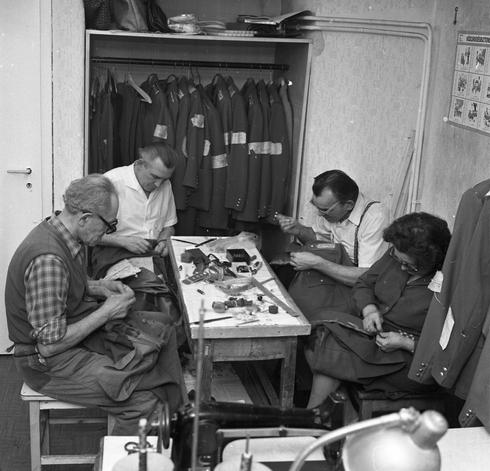 A black and white photo of four people sitting either side of a small, rectangle table, sewing police uniforms. Their heads are bowed as if in prayer. The table is cluttered with various sewing tools and objects such as tape measures rulers, threads. In the background is an open wardrobe where there are Police uniforms on coat hangers.