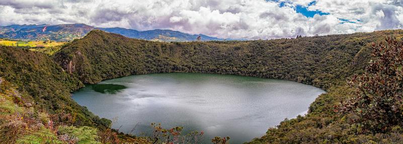 This landscape showcases a lake surrounded by lush greenery in Colombia. The lake's calm waters reflect the sky, creating a serene atmosphere. Nearby mountains and rolling hills provide a backdrop, highlighting the region's natural beauty. Cotton candy clouds float overhead, adding to the picturesque setting.