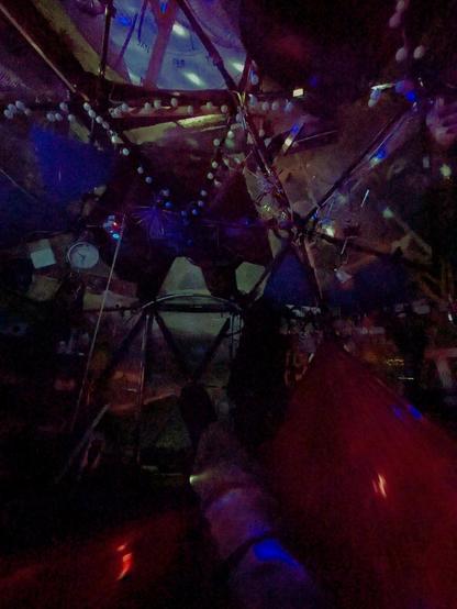 A colorful, cluttered scene of the interior of a room-sized geodesic dome. Christmas lights, tools, and unidentifiable miscellany adorn the space. Booted feet in a hammock are all that is visible of the photographer.