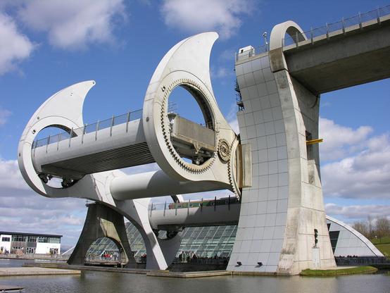 The Falkirk Wheel rotary boat lift. The image shows the Falkirk Wheel from a corner angle with the rotary element halfway through its cycle in the centre of the frame and the support for the fixed end of the canal on the right, with the lower pool bottom left. The visitor centre is in the background. The scene is in sunlight.