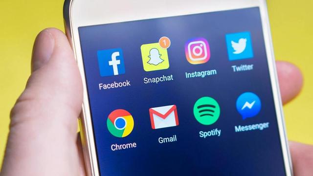 A close-up of a smartphone screen displaying popular app icons, including Facebook, Snapchat, Instagram, Twitter, Chrome, Gmail, Spotify, and Messenger, held in a person's hand.