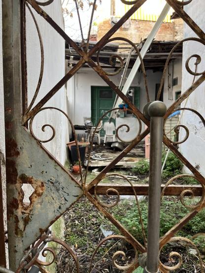 Through a rusted curlicue iron gate, the whitewashed walls of an abandoned courtyard. The paving is green with weeds, the one storey building in the depths has painted green shutters and intact glass in the door but the overhanging roof is falling in and the stacked plastic chairs are grimy, the whole place says neglect.